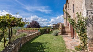 A stone building with a brick pathway leading to a grassy area with a pergola and a view of the mountains.