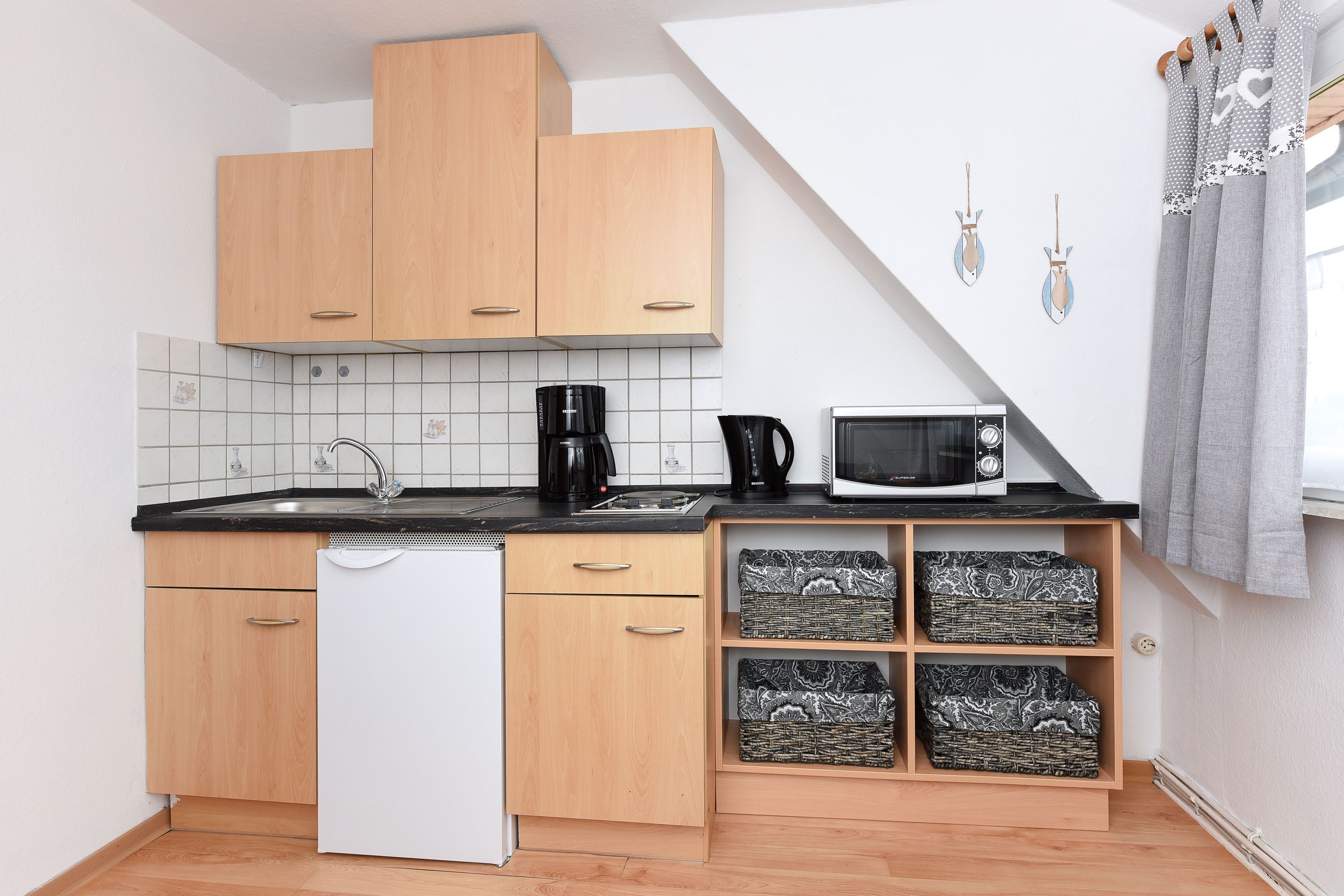 A compact kitchen with wooden cabinets, a white dishwasher, a microwave, and a coffee maker. There are decorative tiles on the backsplash and a small shelf with patterned boxes.