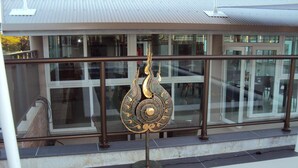 A bronze decorative piece with intricate designs, mounted on a metal stand, placed in front of a building with large windows and a metal roof. - Hotel Colibri (Kone)