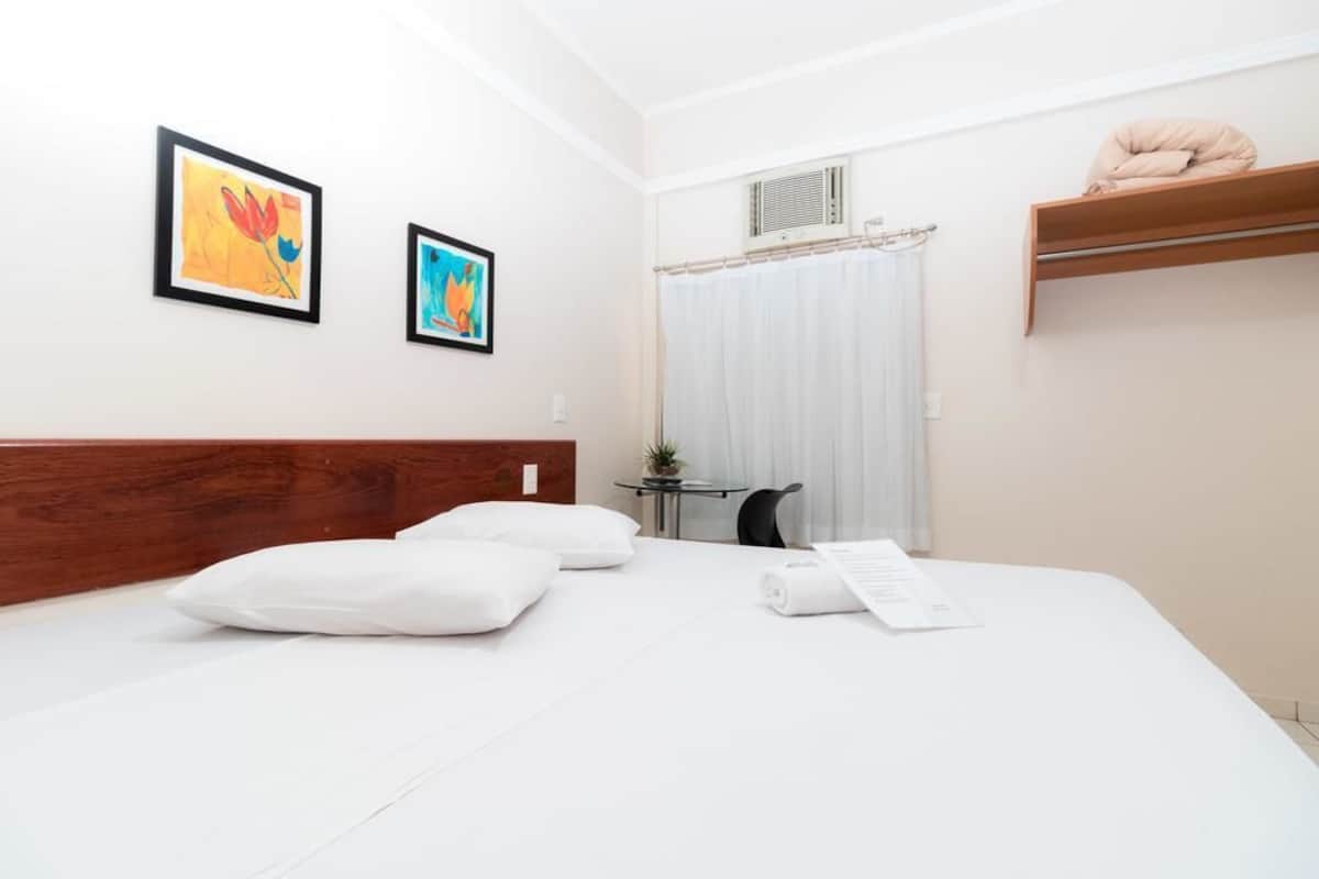 A hotel room with a bed, two pillows, a small table with a plant, and two framed artworks on the wall.