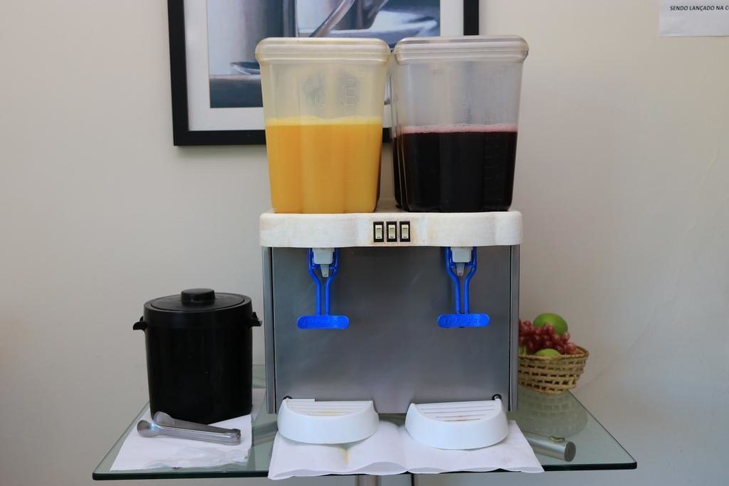 A juice dispenser with two containers, a black container, and a basket of fruit on a table.