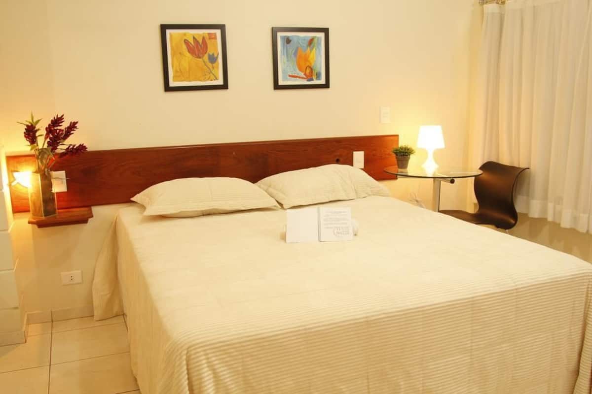 A hotel room with a bed, two framed artworks on the wall, a nightstand with a lamp, and a chair.