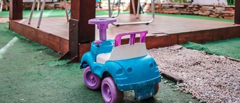 A children's toy car on a green surface with chairs and a wooden deck in the background.