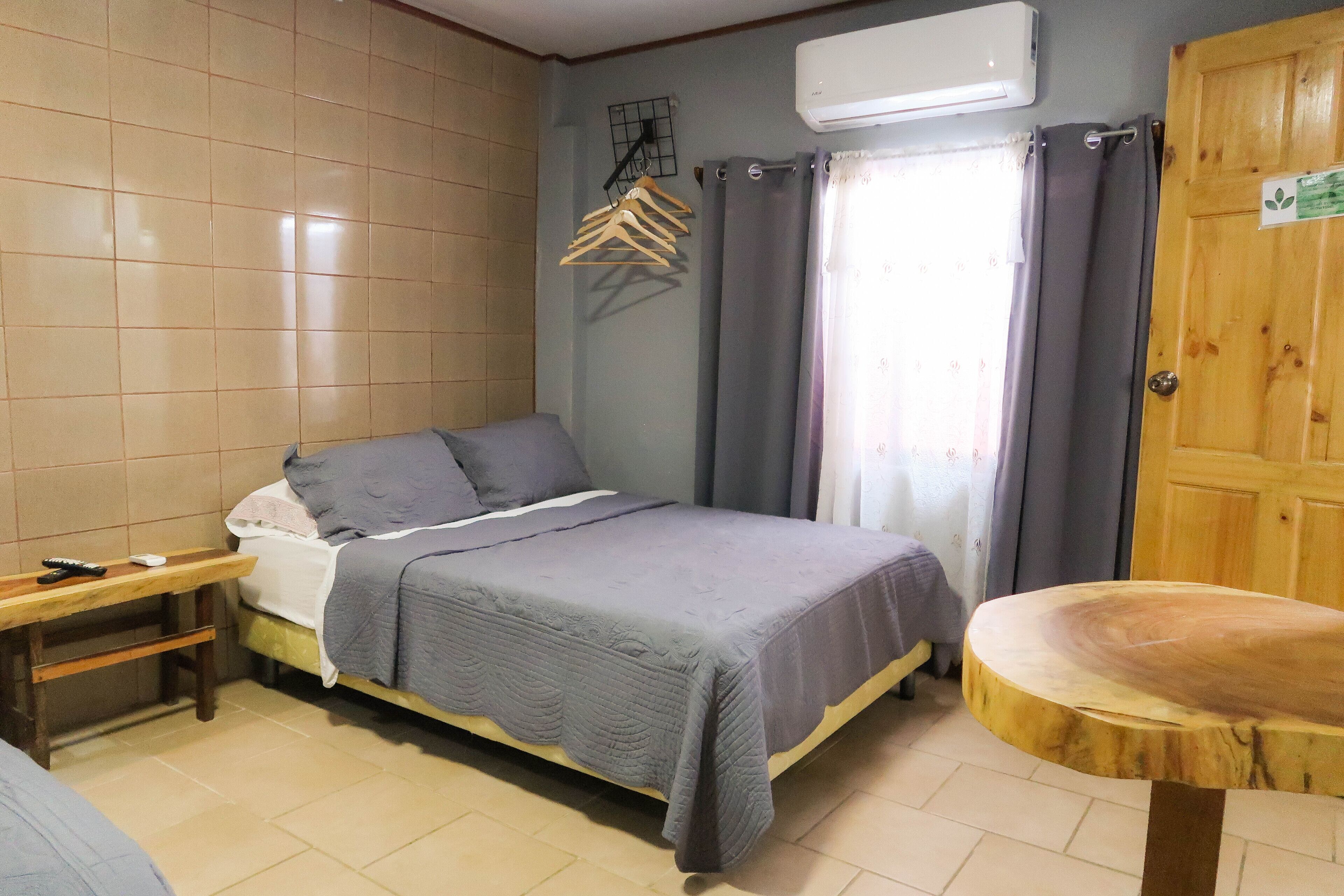 A bedroom with a bed, a wooden table, a hanging clothes rack, and an air conditioning unit.