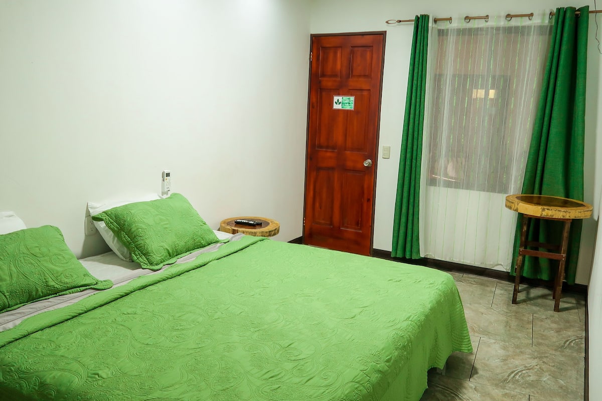 A bedroom with a bed, green bedding, a wooden door, a small round table, and a window with green curtains.