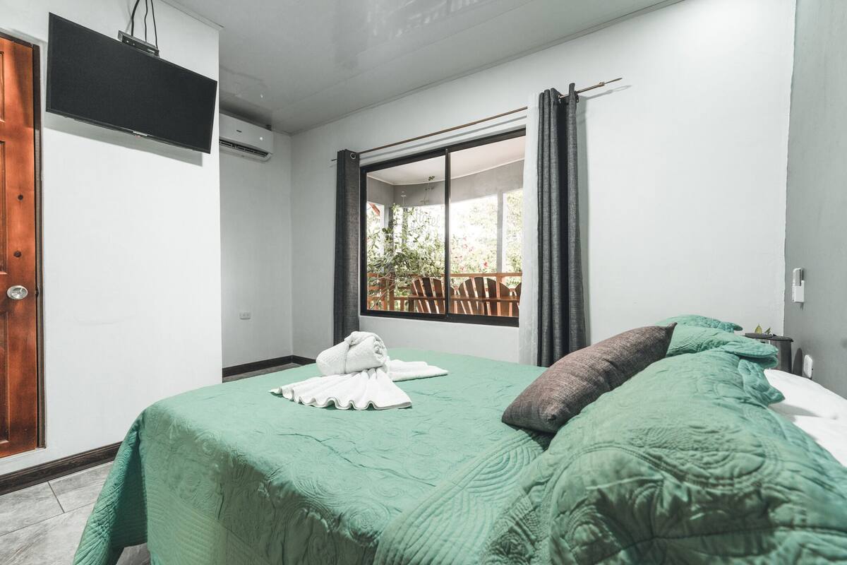 A hotel room with a green bedspread, a flat-screen TV mounted on the wall, and a sliding glass door leading to an outdoor area with a wooden deck.