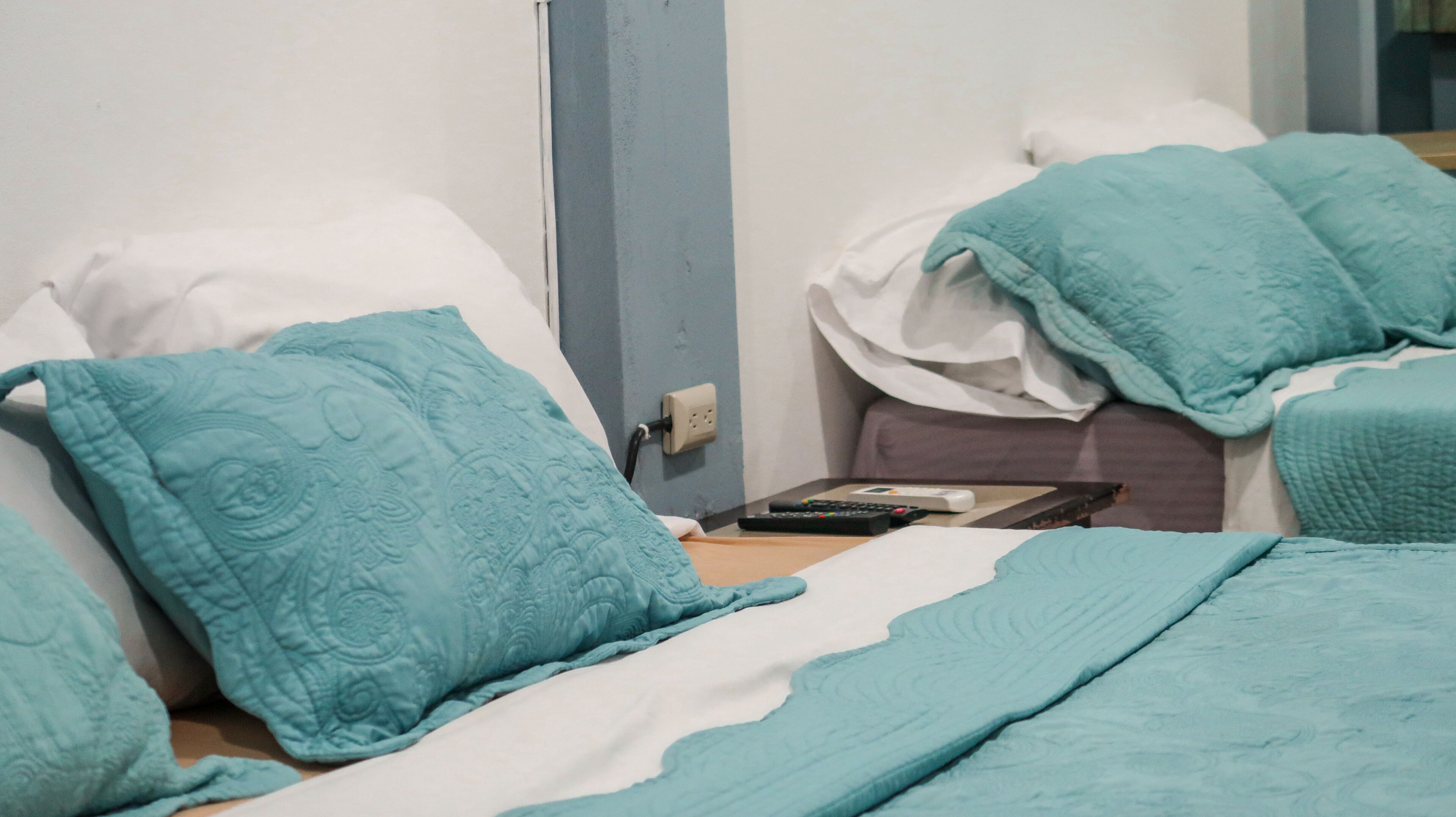A hotel room with two beds, each with a teal quilted pillow and white sheets. A remote control and a book are on the nightstand.