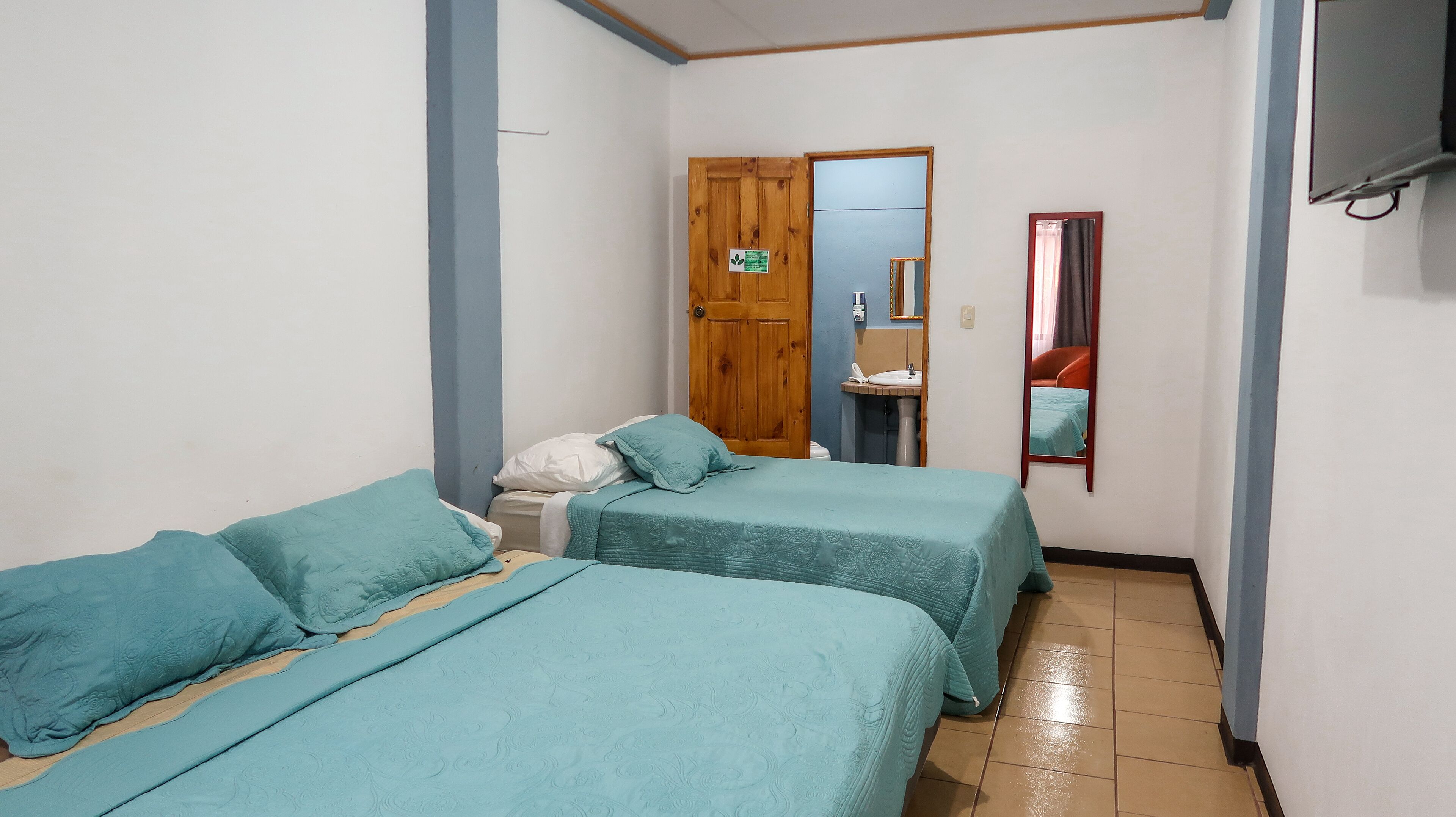 Une petite chambre compacte, avec deux lits simples, une porte en bois, un miroir et une petite table de chevet.