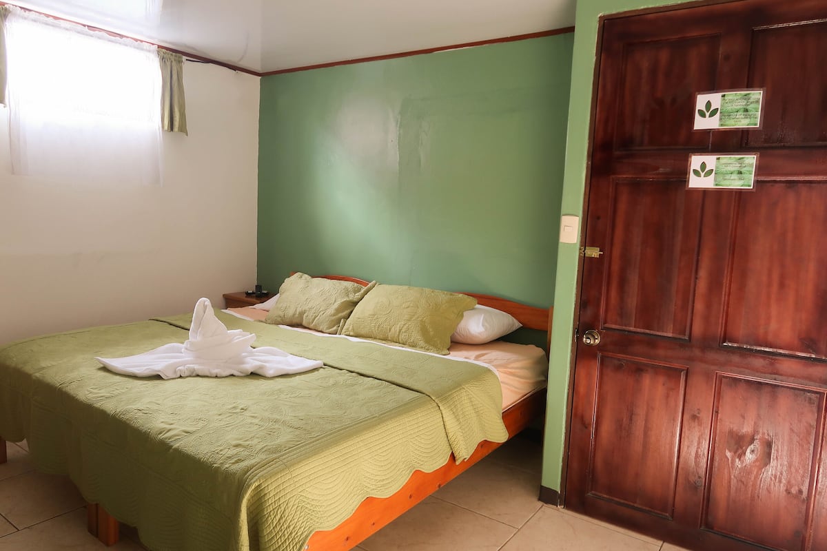 A neatly made bed with a green bedspread and white pillows, a wooden door with a lock, and a window with sheer curtains.