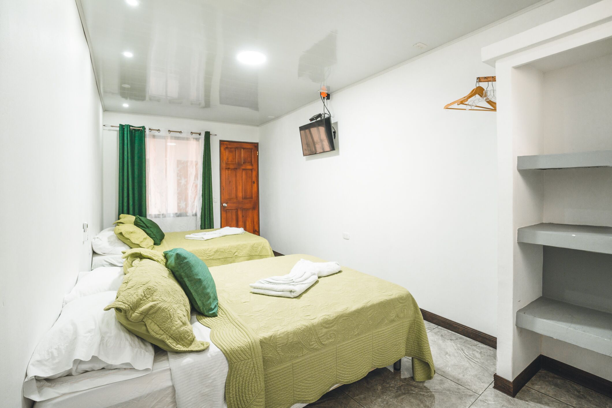 Una habitación limpia y bien iluminada con dos camas, una puerta de madera, un televisor montado en la pared y un perchero.