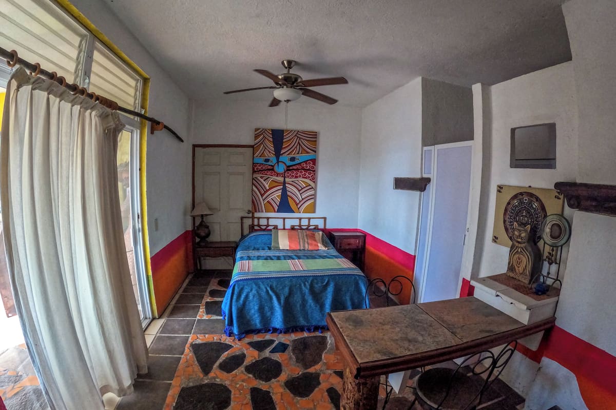 A bedroom with a bed, a ceiling fan, a patterned wall hanging, a small table, and a chair.