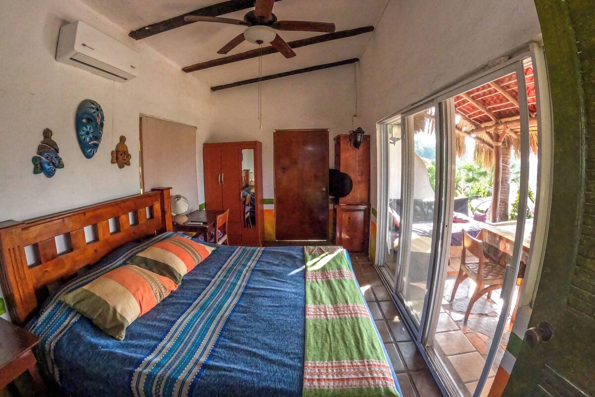 A bedroom with a bed, wooden headboard, ceiling fan, and a sliding glass door leading to an outdoor area with a thatched roof.