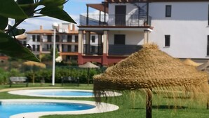 Un área de piscina con una sombrilla de paja, un césped cuidado y un edificio con balcones al fondo.