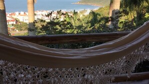 A hammock with a woven seat and a wooden frame, overlooking a coastal landscape with buildings and palm trees. - Pousada Villabella Villaggio (Florianópolis)