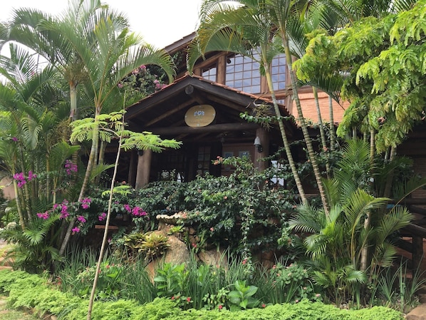 A rustic wooden building with a thatched roof, surrounded by lush tropical vegetation and flowering plants. - Pousada Villabella Villaggio (Florianópolis)