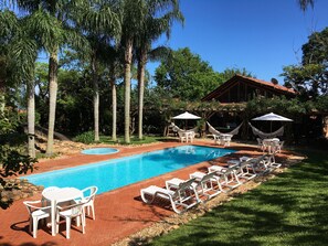 A swimming pool with lounge chairs and umbrellas, surrounded by trees and a building with a red roof. - Pousada Villabella Villaggio (Florianópolis)