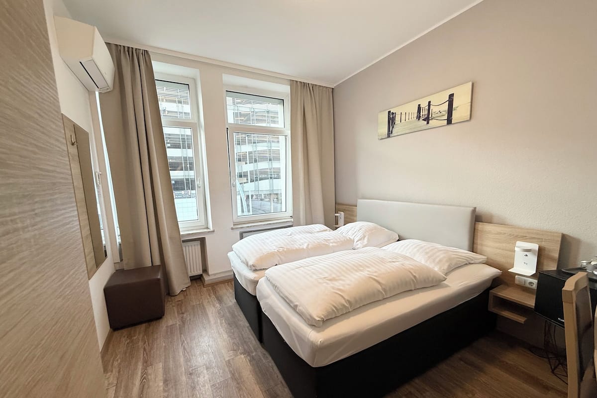 A modern hotel room with a large bed, two windows, a painting on the wall, and a wooden floor.