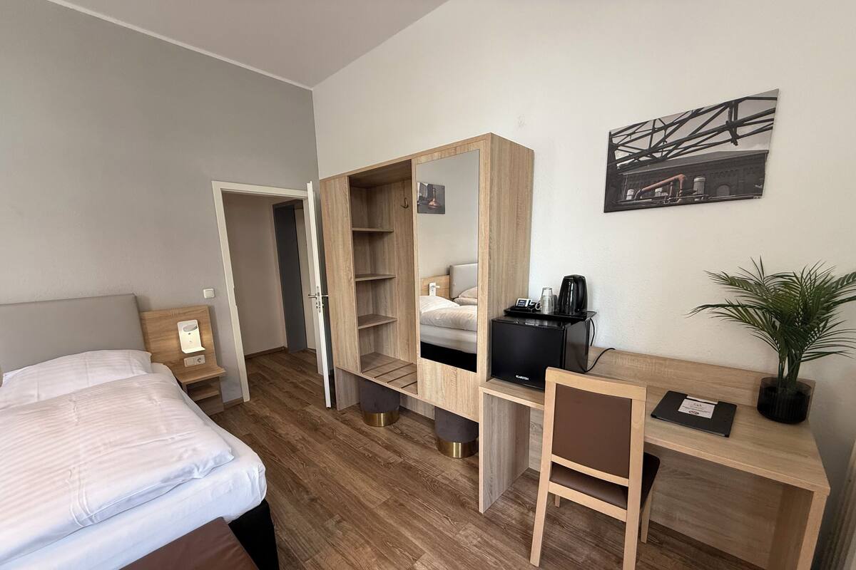 A hotel room with a bed, a desk with a chair, a mirror, a coffee machine, and a plant.