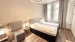 Habitación de hotel moderna con una cama grande, mesitas de noche, un pequeño espacio de descanso y vistas de la ciudad a través de amplias ventanas.