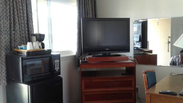A compact hotel room with a television on a wooden stand, a microwave, a small table with a chair, and a window with curtains. - Motel Plaza (Vermilion)