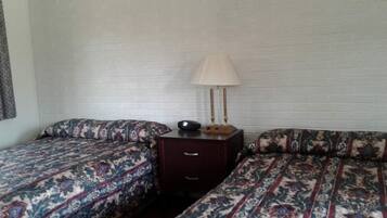 A room with two beds, each with patterned bedding, a bedside table with a lamp, and a small drawer.