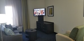 A hotel room with a flat-screen TV mounted on the wall, a sofa, a small round table, and a cabinet. - Candlewood Suites Plano North by IHG (Plano)