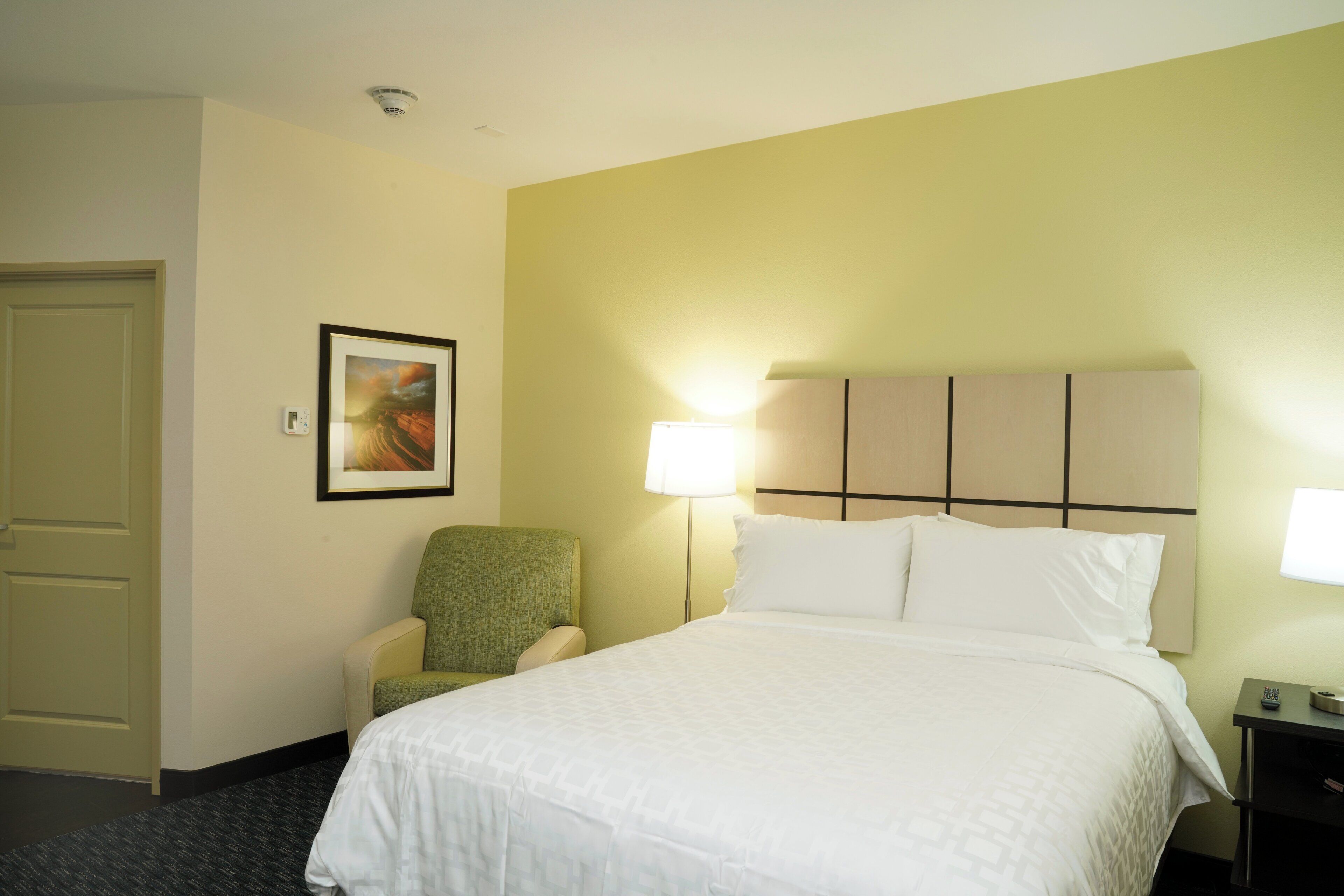 A hotel room with a bed, a chair, a wall-mounted picture, and a bedside table with a lamp.