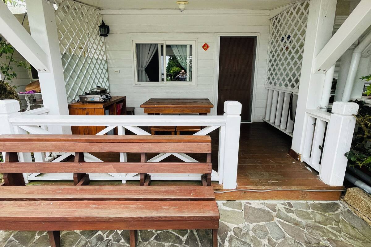 A covered porch with wooden benches and tables, a stone floor, and a white fence.