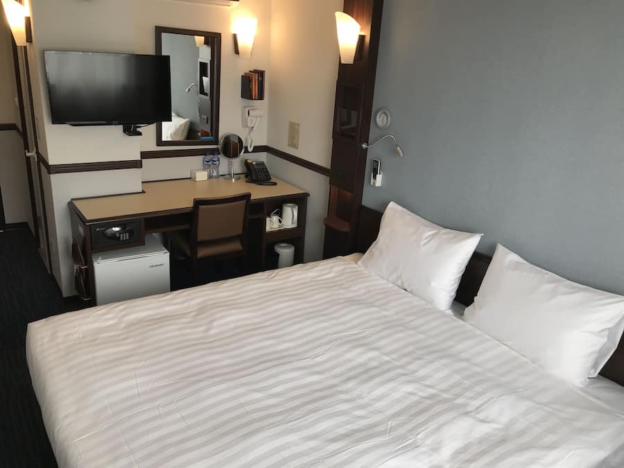 Una habitación de hotel con cama, escritorio, silla, televisor, espejo, teléfono y minibar.