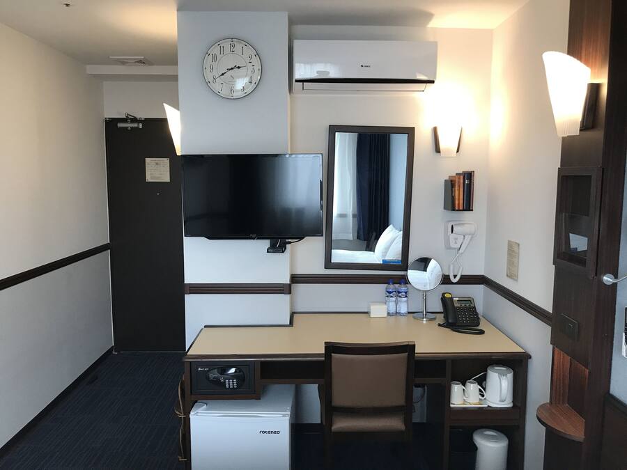Habitación de hotel con escritorio, silla, minibar, reloj, televisor, espejo y puerta con manija.