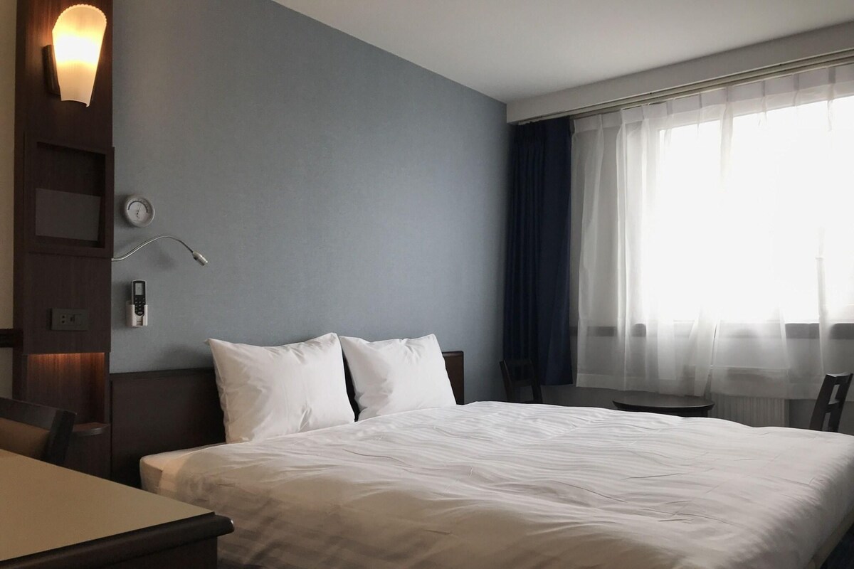 A hotel room with a bed, a desk, a chair, a clock, and a window with curtains.