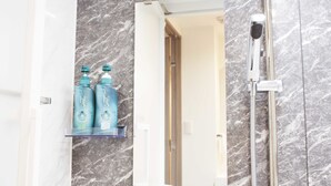 Un baño con pared de mármol, repisa de vidrio con artículos de aseo y ducha con cabezal de ducha de mano.