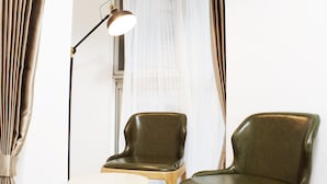 Una habitación con dos sillones de cuero verde y una mesita redonda pequeña, una lámpara de pie y un ventanal con cortinas traslúcidas.