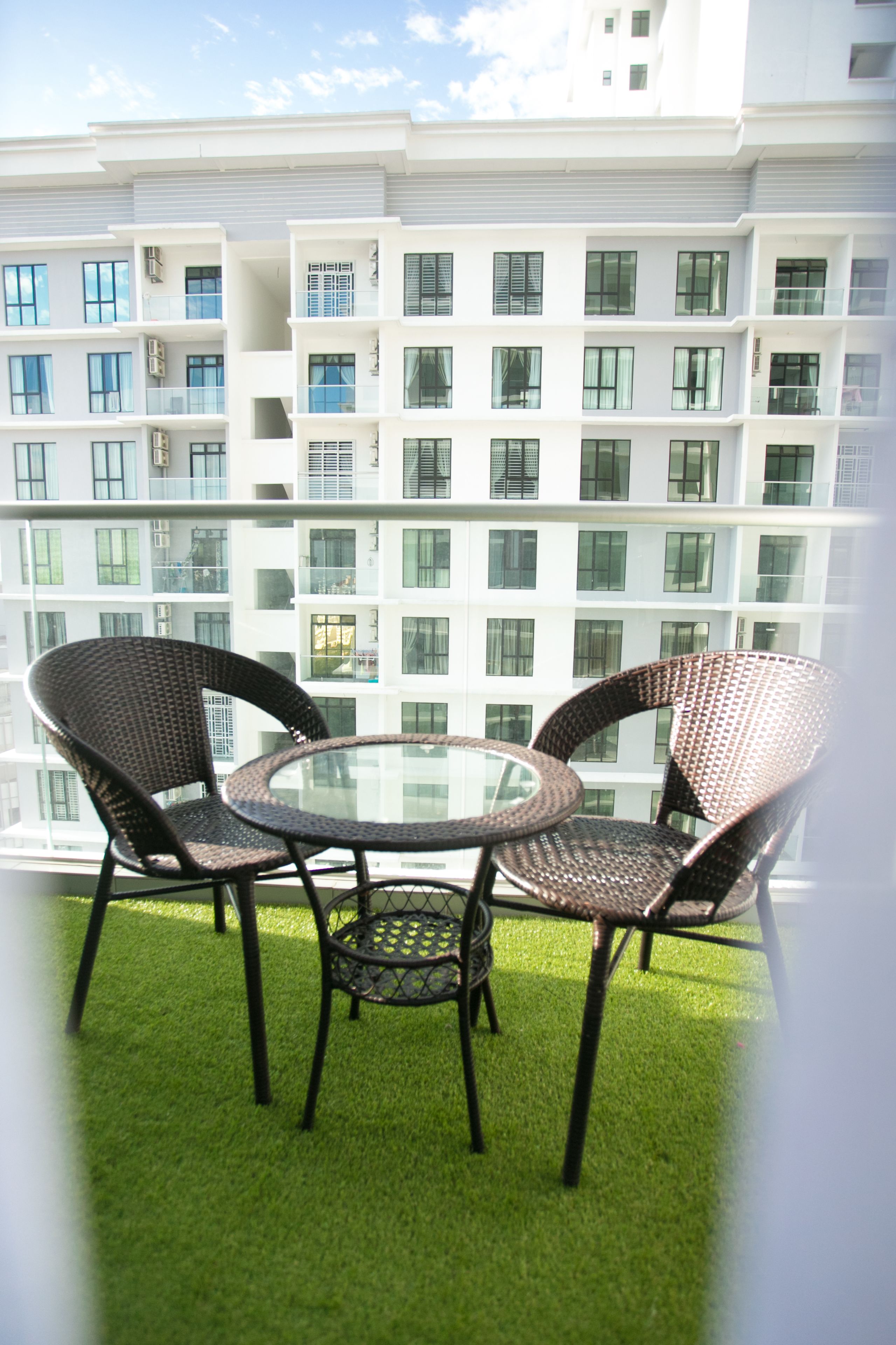 Un balcone con due poltrone in vimini e un tavolino in vetro, con vista su un edificio moderno dotato di numerosi balconi.