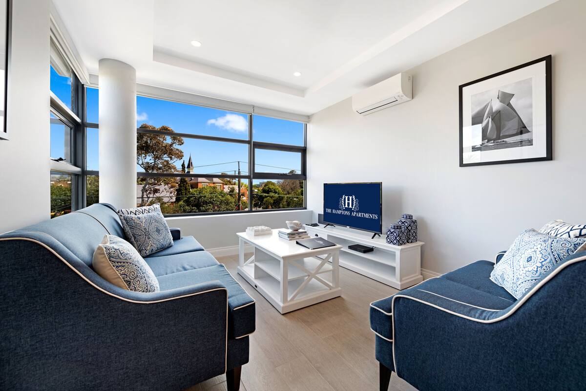 A modern living room with a sofa, coffee table, and TV, large windows with a view of trees and buildings, and a framed picture on the wall.