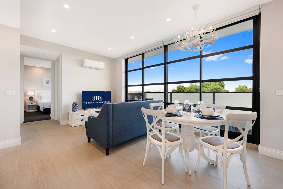 A modern living room with a large window, a dining table with white chairs, a blue sofa, and a television.