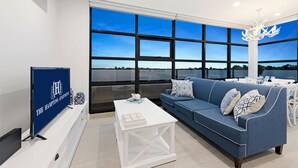 Een moderne woonkamer met een groot raam, een blauwe bank, een witte salontafel en een flatscreen televisie op een voet.