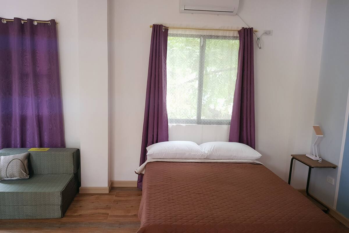 A room with a bed, a small couch, a window with purple curtains, and a bedside table with a lamp.