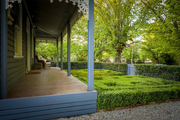 A porch with a covered seating area, a hedge, and a tree in the background.