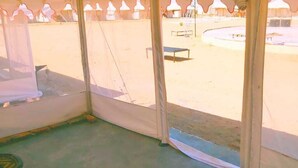 A room with a large window overlooking a courtyard with a pool, outdoor furniture, and thatched-roof structures. - Desert Raasleela Camp (Jaisalmer)