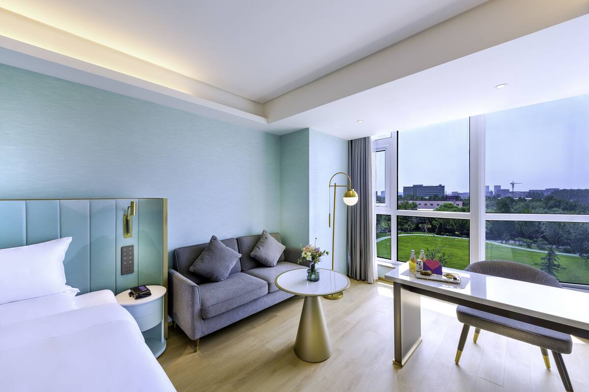 Ein modernes Hotelzimmer mit Bett, Sofa, rundem Tisch und Blick auf eine Stadtlandschaft durch große Fenster.