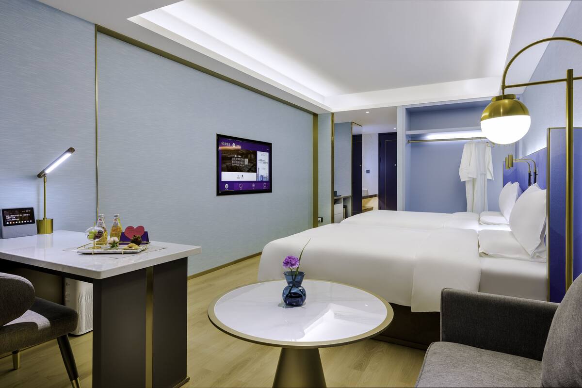 Ein modernes Hotelzimmer mit einem Bett, einem Schreibtisch mit Computer, einem runden Tisch mit Vase und einem an der Wand befestigten Fernseher.