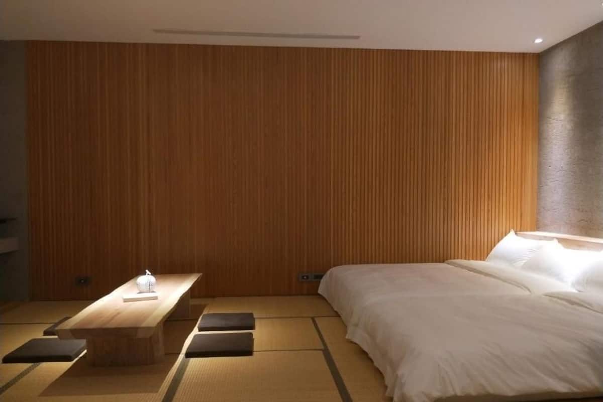 A modern hotel room with a wooden panel wall, a bed with white bedding, a wooden table with a lamp, and tatami mats on the floor.
