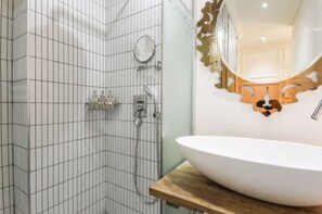 A bathroom with a white tiled shower, a round mirror with a decorative frame, and a large white sink on a wooden countertop.