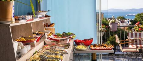 A buffet with various food items and utensils, a potted plant, and a view of the sea and buildings.