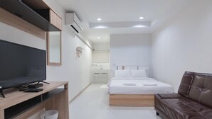 A modern hotel room with a wooden desk, a flat-screen TV, a bed with white linens, a leather sofa, and a small bathroom with a sink and mirror. - The Chill House (Tha Muang)
