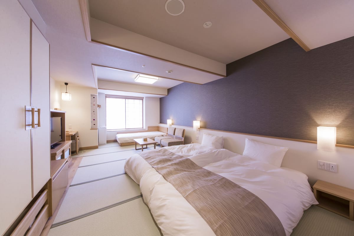 A hotel room with a large bed, a sitting area with a sofa and a small table, a television, and a window with natural light.