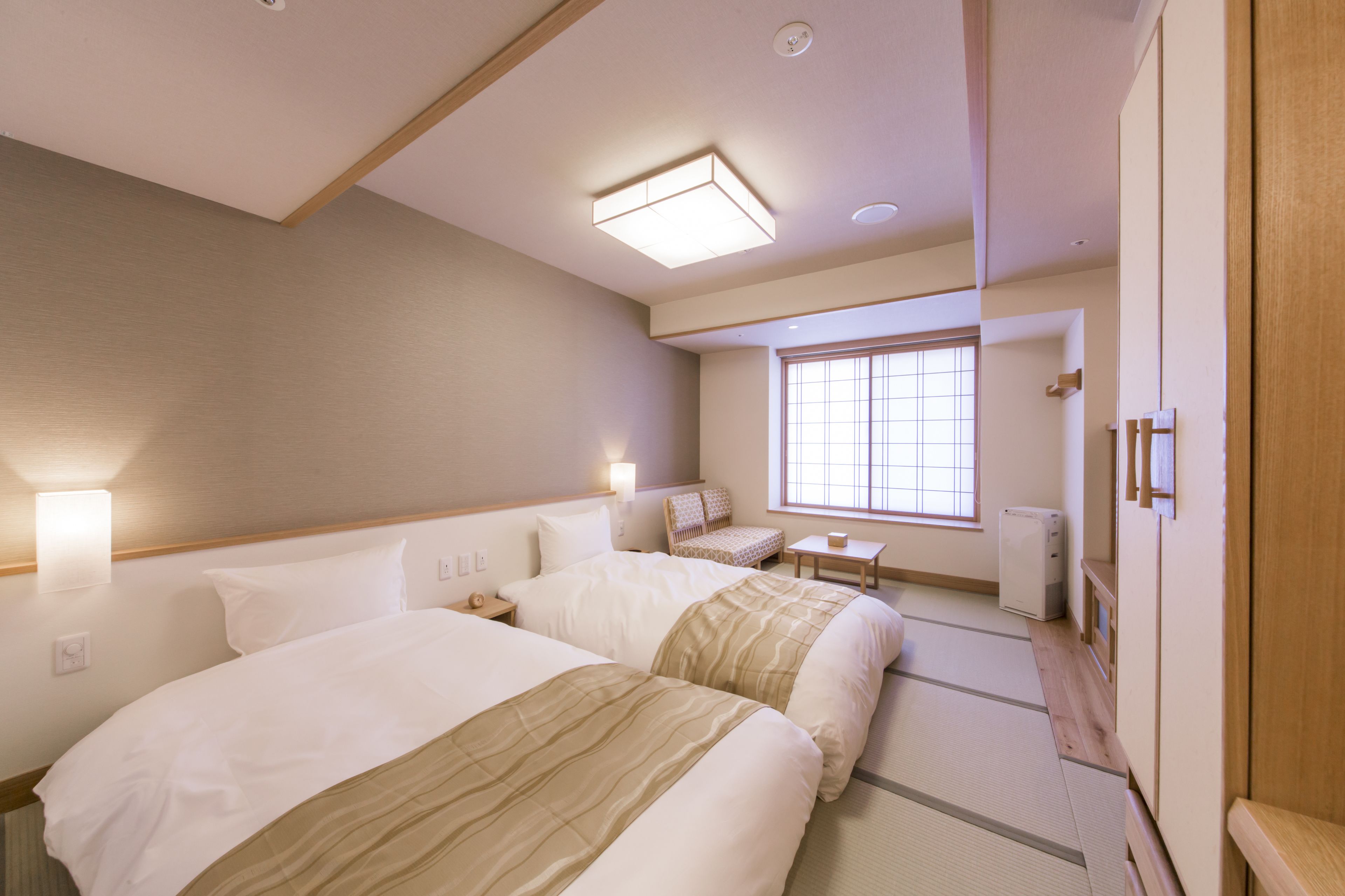A hotel room with two beds, a small table, a chair, a window, and a wardrobe.