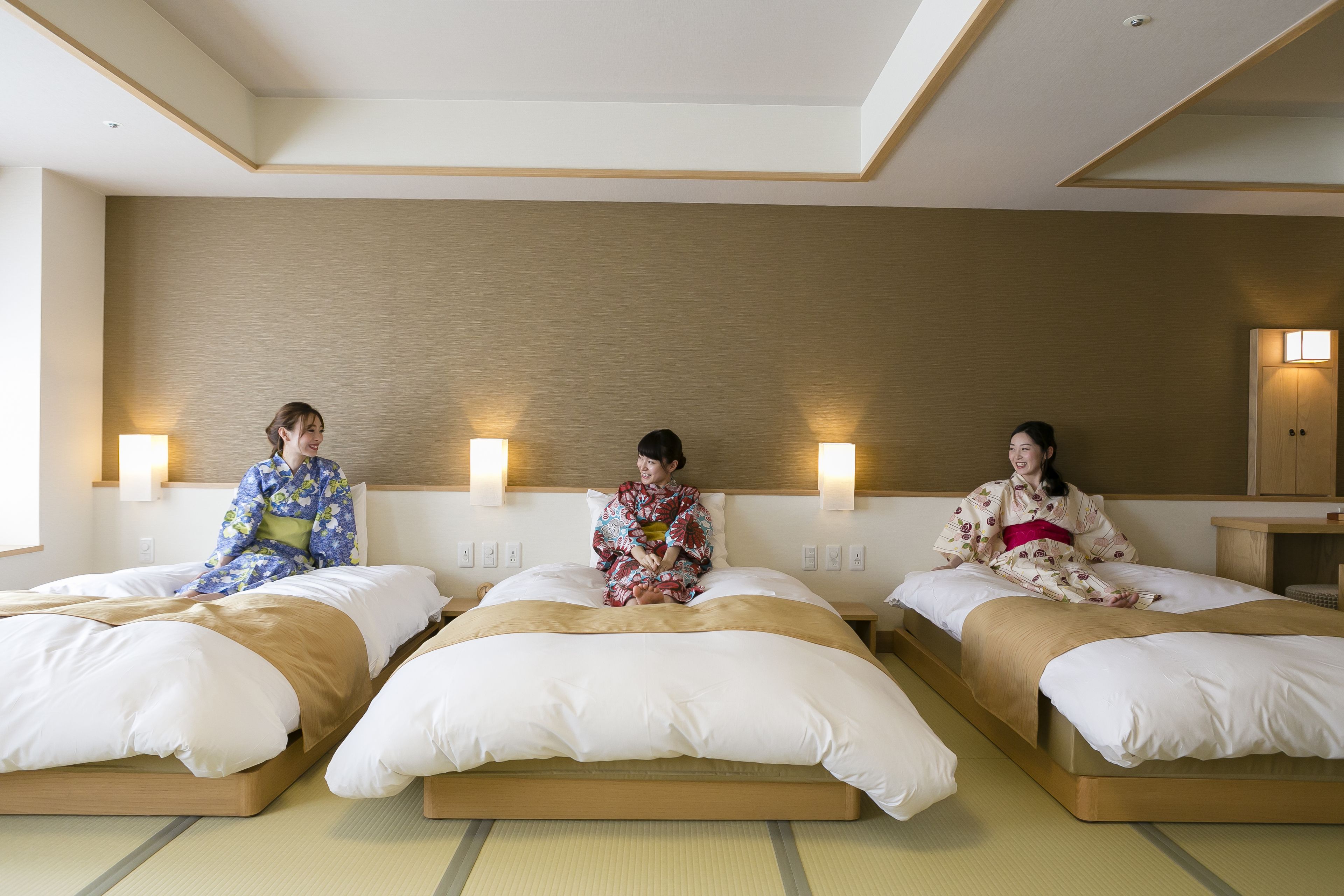 Three beds in a hotel room with a wooden headboard, a wall-mounted light fixture, and a small wooden nightstand.