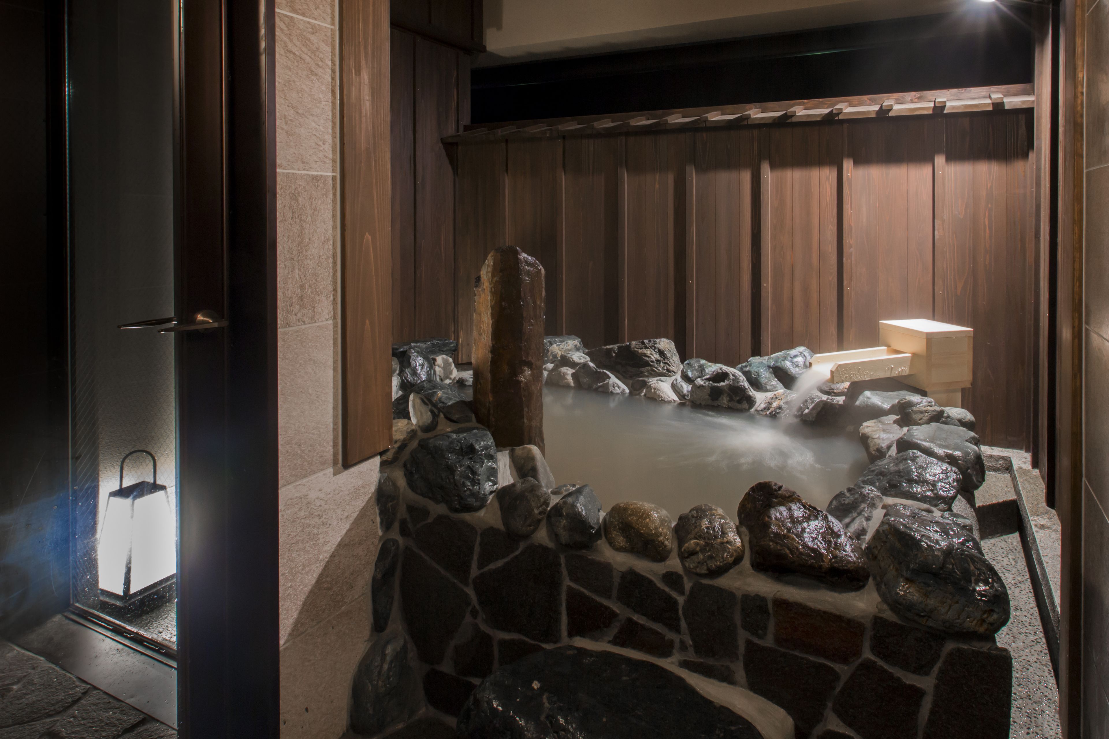 A stone hot tub with a wooden backdrop and a stone wall surrounding it.